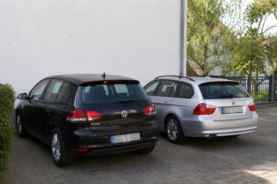 Stellplatz zu vermieten Zwei geparkte Autos stehen nebeneinander auf einem gepflasterten Parkplatz vor der weißen Außenwand eines Wohngebäudes. Links ist ein dunkler Kompaktwagen, rechts ein silberner Kombi zu sehen. Im Hintergrund befinden sich Bäume und ein Zaun. Die Stellplätze wirken gepflegt und befinden sich in einer ruhigen Wohnumgebung. Die Kennzeichen der Fahrzeuge sind unkenntlich gemacht.