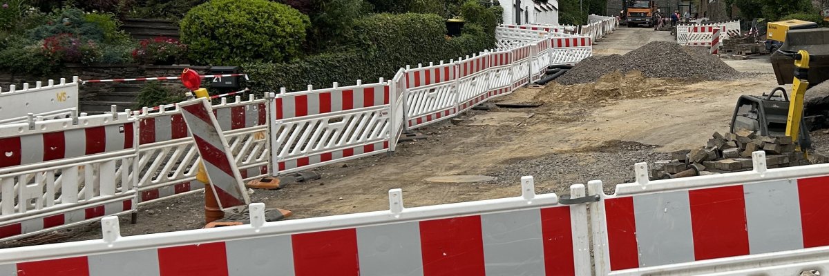Mehrere rot-weiße Absperrbaken sichern eine Straßenbaustelle. Im Hintergrund sind eine Straße, ein Hang mit Bäumen und weitere Baustelleneinrichtungen zu sehen.
