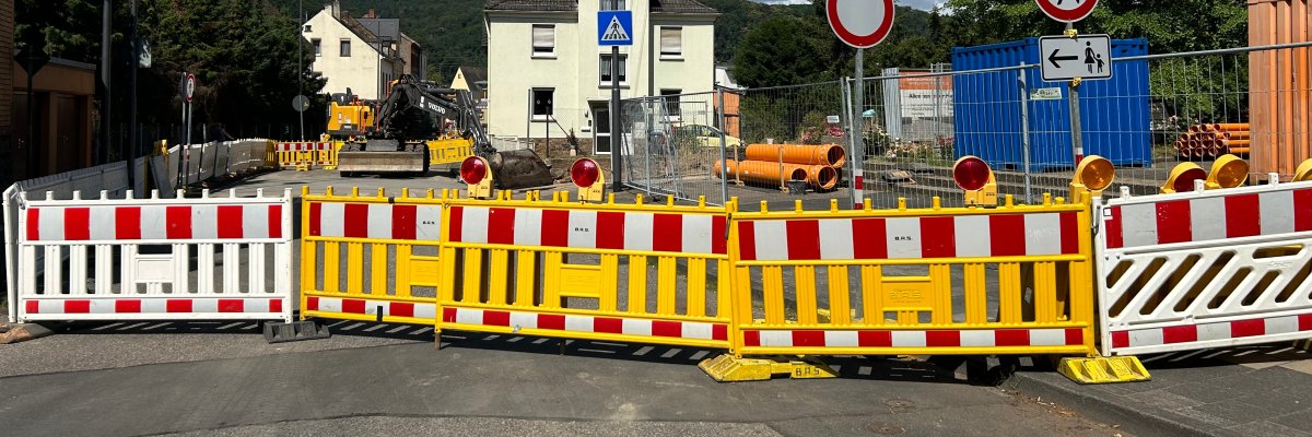 Baustelle Johann-Baptist-Ludwig-Straße Lahnstein Eine vollständig abgesperrte Straße, die Johann-Baptist-Ludwig-Straße, mit rot-weißen und gelben Baustellenbaken. Im Hintergrund sind Wohnhäuser, Bäume und bewaldete Hänge zu sehen.