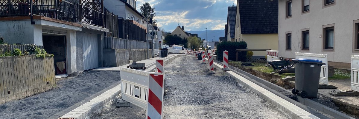 Asphaltarbeiten Hohenrhein Die Straße Hohenrhein in Lahnstein, deren Asphaltdecke abgetragen ist. Links und rechts sind Häuserreihen zu sehen sowie vereinzelte rot-weiß gestreifte Absperrpoller. Der blaue Himmel ist wolkenverhangen.