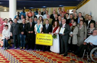 Eine große Gruppe von rund 40 Personen steht in einem Veranstaltungsgebäude, der Stadthalle Lahnstein. Die Menschen stehen in mehreren Reihen für ein Erinnerungsfoto zusammen. In der ersten Reihe wird ein Schild mit der Aufschrift „Fairtrade-Stadt Lahnstein“ präsentiert. Einige Personen halten Urkunden oder Blumen. Der Innenraum ist durch einen auffällig gemusterten Teppich und eine blau gestaltete Decke geprägt.