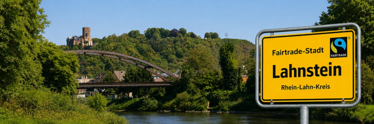 Panoramaaufnahme von Lahnstein mit Blick auf die Lahn und die Burg Lahneck auf dem bewaldeten Hügel im Hintergrund. Im Vordergrund steht links ein großes gelbes Ortsschild mit der Aufschrift „Fairtrade-Stadt Lahnstein“ und Fairtrade-Logo. Eine Brücke überspannt die Lahn, umgeben von üppigem Grün und blauem Himmel. Das Bild dient als Headergrafik für die Fairtrade-Stadt Lahnstein.