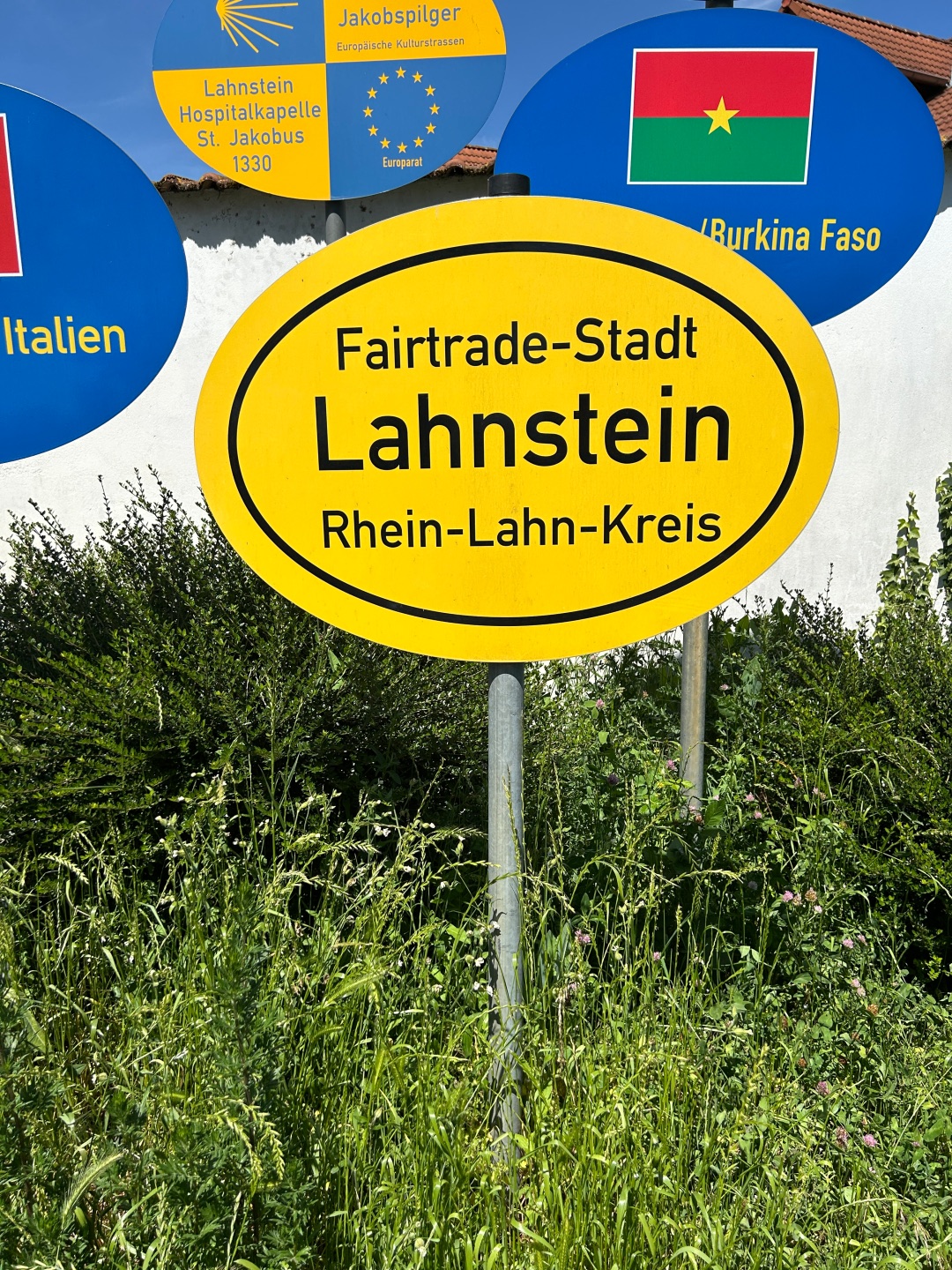 Detailaufnahme des gelben Schildes „Fairtrade-Stadt Lahnstein / Rhein-Lahn-Kreis“. Das Schild nimmt den größten Teil des Bildes ein und steht auf einem Metallpfosten. Dahinter sind Teile weiterer Partnerschafts- und Europaschilder zu sehen. Im Vordergrund wächst hohes Gras mit Wildblumen.