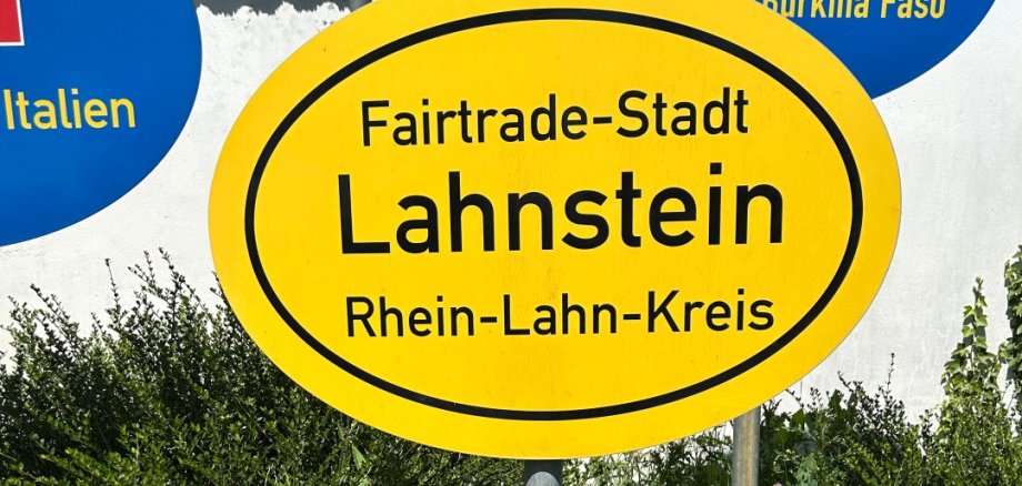 Fairtrade-Stadt Lahnstein Detailaufnahme des gelben Schildes „Fairtrade-Stadt Lahnstein / Rhein-Lahn-Kreis“. Das Schild nimmt den größten Teil des Bildes ein und steht auf einem Metallpfosten. Dahinter sind Teile weiterer Partnerschafts- und Europaschilder zu sehen. Im Vordergrund wächst hohes Gras mit Wildblumen.