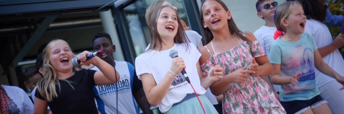 Mehrere Kinder und Jugendliche stehen mit Mikrofonen auf einer Bühne und singen gemeinsam. Im Vordergrund sind zwei Solistinnen zu sehen, im Hintergrund begleitet eine Gruppe den Auftritt.