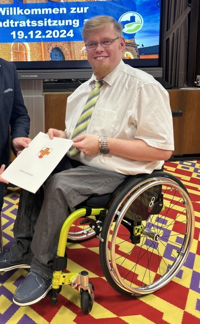 Ein lächelnder Mann mit Brille und heller Krawatte, der Behindertenbeauftragte der Stadt Lahnstein, Dennis Bloem, sitzt in einem Rollstuhl in einem Sitzungssaal. Im Hintergrund ist ein Bildschirm mit der Begrüßung zur Stadtratssitzung zu sehen, auf dem "Willkommen zur Stadtratssitzung am 19.12.2024" steht.