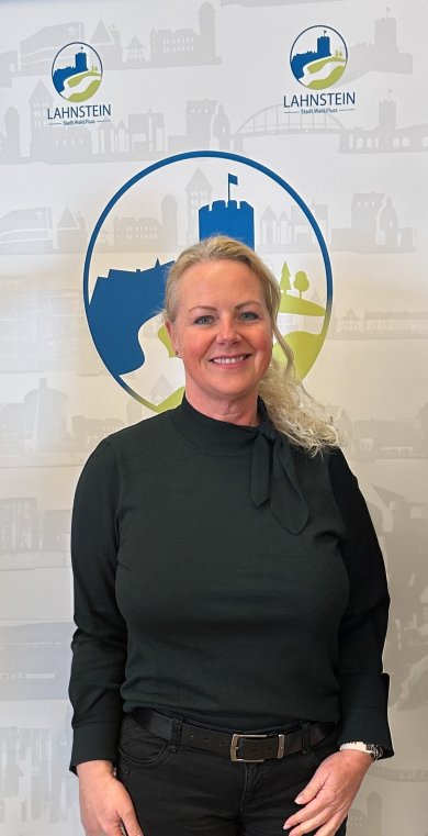 Porträt einer lächelnden Frau mit zusammengebundenen Haaren vor einer hellen Wand. Im Hintergrund ist ein Teil des Logos der Stadt Lahnstein zu erkennen. Es handelt sich um die Gleichstellungsbeauftragte der Stadt Lahnstein Tatjana Schmitt.