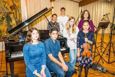Kreismusikschule Rhein-Lahn Eine Gruppe von sieben Kindern und zwei Erwachsenen steht in einem Musikraum um einen Flügel herum. Einige der Kinder halten Instrumente, darunter eine Querflöte und eine Geige. Im Hintergrund sind Mikrofone und Notenständer aufgebaut, was auf eine musikalische Aufnahme oder Probe hindeutet. Die Gruppe blickt in die Kamera und wirkt freundlich und konzentriert.