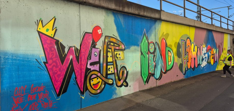 Ein farbenfrohes, großflächiges Graffiti-Wandbild auf einer Betonmauer unterhalb einer Brücke, das den Schriftzug „WIR SIND LAHNSTEIN“ in verschiedenen künstlerischen Stilen und bunten Farben zeigt. Rechts steht eine Person mit Warnweste und blickt zur Wand. Unten links sind einige Namen gesprüht.