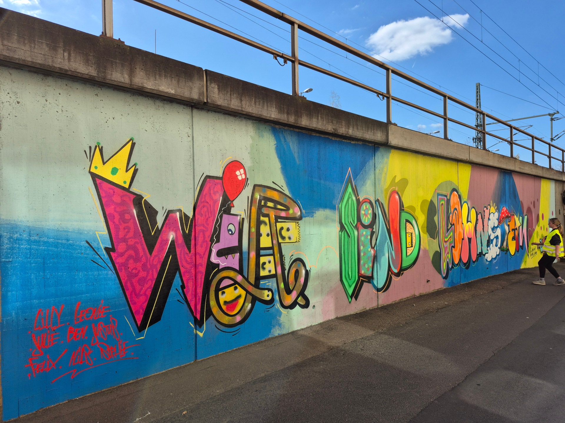 Ein farbenfrohes, großflächiges Graffiti-Wandbild auf einer Betonmauer unterhalb einer Brücke, das den Schriftzug „WIR SIND LAHNSTEIN“ in verschiedenen künstlerischen Stilen und bunten Farben zeigt. Rechts steht eine Person mit Warnweste und blickt zur Wand. Unten links sind einige Namen gesprüht.