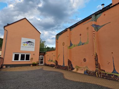 Außenansicht des Jugendkulturzentrums Lahnsteins mit apricotfarbenen Gebäudewänden, die teils von Fliesen im Hundertwasserstil bedeckt sind, unter einem bewölkten Himmel. An der Hauswand hängt ein großes weißes Schild, auf dem das JUKZ-Logo abgebildet ist und in schwarz steht: JUKZ Lahnstein Haus der Generationen"
