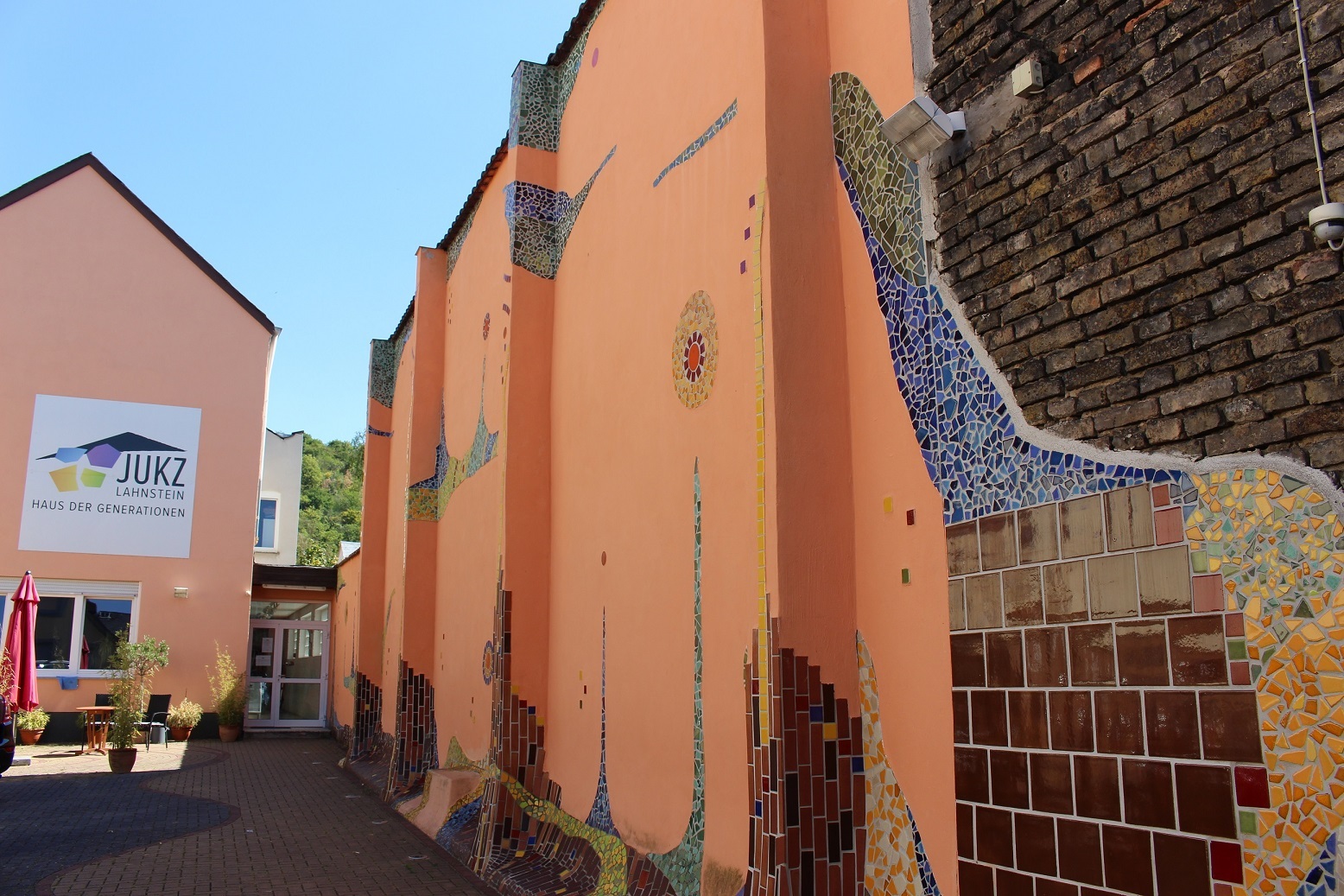 Blick in den Hof des Jugendkulturzentrums Lahnstein mit Pflanzen sowie Blick auf ein apricotfarbenes Gebäude. Die Wänder der Mauer rechts sind teils von Fliesen im Hundertwasserstil bedeckt; blauer Himmel ist erkennbar. An der Hauswand hängt ein großes weißes Schild, auf dem das JUKZ-Logo abgebildet ist und in schwarz steht: JUKZ Lahnstein Haus der Generationen"