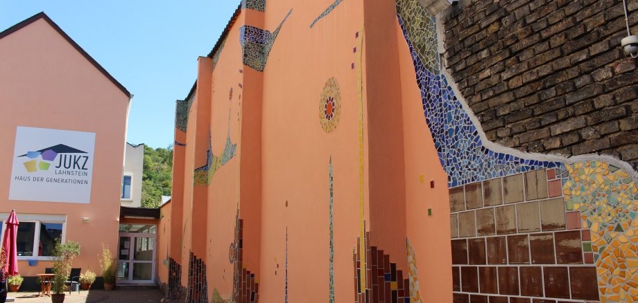 Jugendkulturzentrum Lahnstein Blick in den Hof des Jugendkulturzentrums Lahnstein mit Pflanzen sowie Blick auf ein apricotfarbenes Gebäude. Die Wänder der Mauer rechts sind teils von Fliesen im Hundertwasserstil bedeckt; blauer Himmel ist erkennbar. An der Hauswand hängt ein großes weißes Schild, auf dem das JUKZ-Logo abgebildet ist und in schwarz steht: JUKZ Lahnstein Haus der Generationen"