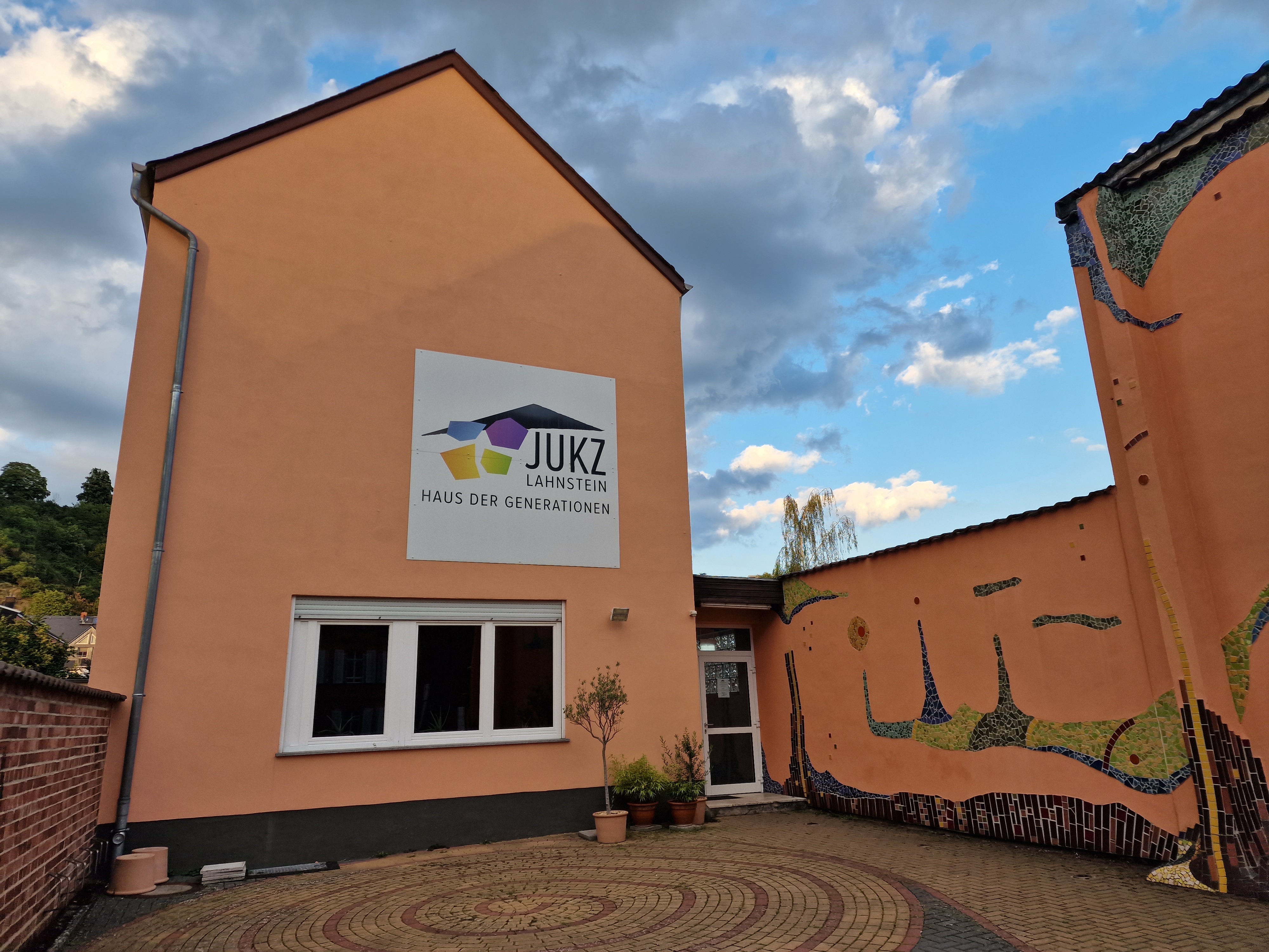 Außenansicht des Jugendkulturzentrums Lahnsteins mit apricotfarbenen Gebäudewändenm, die teils von Fliesen im Hundertwasserstil bedeckt sind, unter einem bewölkten Himmel. An der Hauswand hängt ein großes weißes Schild, auf dem das JUKZ-Logo abgebildet ist und in schwarz steht: JUKZ Lahnstein Haus der Generationen"