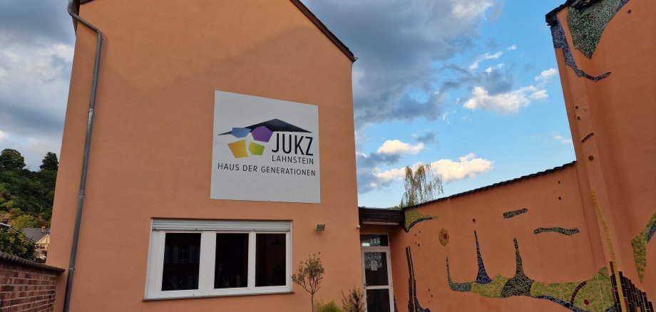Jugendkulturzentrum Lahnstein Außenansicht des Jugendkulturzentrums Lahnsteins mit apricotfarbenen Gebäudewändenm, die teils von Fliesen im Hundertwasserstil bedeckt sind, unter einem bewölkten Himmel. An der Hauswand hängt ein großes weißes Schild, auf dem das JUKZ-Logo abgebildet ist und in schwarz steht: JUKZ Lahnstein Haus der Generationen"