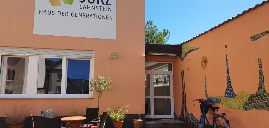 Jugendkulturzentrum Lahnstein Der Eingangsbereich des Jugendkulturzentrums mit einer kleinen Außenterrasse, Tisch und Stühlen. Das Gebäude ist apricotfarbenen, die Mauer im rechten Bildteils ist teils von Fliesen im Hundertwasserstil bedeckt; es herrscht ein bewölkter Himmel. An der Hauswand hängt ein großes weißes Schild, auf dem das JUKZ-Logo abgebildet ist und in schwarz steht: JUKZ Lahnstein Haus der Generationen".