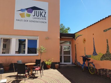 Jugendkulturzentrum Lahnstein Der Eingangsbereich des Jugendkulturzentrums mit einer kleinen Außenterrasse, Tisch und Stühlen. Das Gebäude ist apricotfarbenen, die Mauer im rechten Bildteils ist teils von Fliesen im Hundertwasserstil bedeckt; es herrscht ein bewölkter Himmel. An der Hauswand hängt ein großes weißes Schild, auf dem das JUKZ-Logo abgebildet ist und in schwarz steht: JUKZ Lahnstein Haus der Generationen".