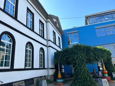 Blick auf den Hof mit einem historischen, weiß verputzten Gebäude und einem modernen Anbau mit blauer Fassade. Eine mit Efeu bewachsene Pergola verbindet die beiden Gebäudeteile. Unter der Pergola stehen mehrere farbenfrohe Holzfiguren oder Spielobjekte. Der Himmel ist klar und blau.
