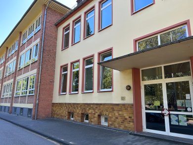 Mehrgeschossiges Schulgebäude mit einer hell verputzten Fassade und einem angrenzenden Backsteinbau. Rechts befindet sich ein überdachter Eingangsbereich mit verglasten Türen. Die Fassade ist durch regelmäßig angeordnete Fenster gegliedert, vor dem Gebäude verläuft ein Gehweg.