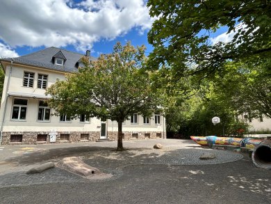 Schulhof mit einem hell verputzten, mehrgeschossigen Schulgebäude. In der Mitte steht ein großer Laubbaum, rechts befinden sich ein Basketballkorb sowie eine farbenfrohe Spiel- und Sitzskulptur. Der Hof ist mit Pflasterflächen und Spielobjekten gestaltet.
