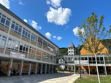 Moderner Schulcampus des Privaten Johannes-Gymnasiums Lahnstein mit mehreren Gebäudeteilen, einem überdachten Verbindungsgang und einem offenen Schulhof. Im Hintergrund ragt der Turm einer Kirche vor bewaldeten Hügeln auf.