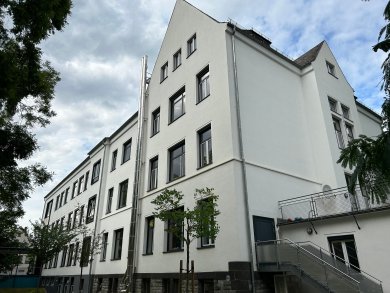 Seitliche Ansicht eines mehrstöckigen Gebäudes mit weißer Fassade, zahlreichen Fenstern und modernisiertem Eingangsbereich. Kleine Bäume und Grünflächen säumen den Weg entlang des Gebäudes, darüber ein leicht bewölkter Himmel.
