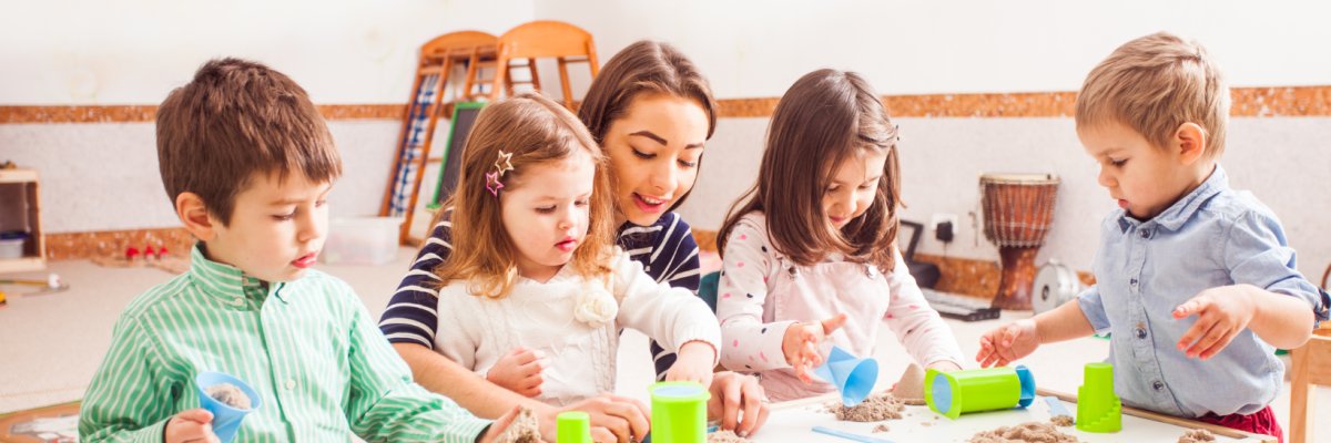 Eine Frau begleitet vier Kinder beim gemeinsamen Spielen mit kinetischem Sand an zwei Spieltischen. Die Kinder formen und schütten den Sand mit Bechern und Förmchen und entdecken spielerisch verschiedene Materialien in einem hellen Gruppenraum.