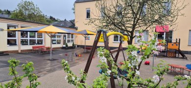 Blick auf das großzügige Außengelände einer Kindertagesstätte mit Sandspielbereich, Schaukeln, Klettergerüst und Sonnenschirmen. Sitzgelegenheiten und Spielgeräte laden zum Spielen und Verweilen im Freien ein.