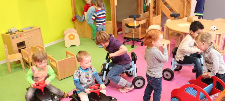 Kita EinSteinchen Mehrere Kinder nutzen unterschiedliche Spielbereiche eines hellen Gruppenraums. Einige fahren mit Rutschfahrzeugen über den farbigen Boden, andere spielen an kleinen Tischen, in einer Spielhöhle oder beschäftigen sich mit Rollenspielen. Der Raum ist mit altersgerechten Möbeln und vielfältigen Spielmaterialien ausgestattet und lädt zum freien Spielen ein.