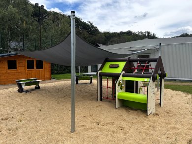 Kita Kastanienplatz am Ersatzstandort in Friedrichssegen Außengelände der Kita Kastanienplatz am Ersatzstandort in Friedrichssegen. Auf einer großen Sandfläche steht ein grün-schwarzes Spielgerät mit Kletternetzen und kleiner Spieltheke. Ein großflächiges dunkles Sonnensegel überspannt einen Teil des Spielbereichs. Links befindet sich ein Holzhaus, außerdem stehen mehrere Sitzbänke auf der Fläche. Im Hintergrund sind das Kita-Gebäude sowie Bäume und bewachsene Hänge zu sehen.
