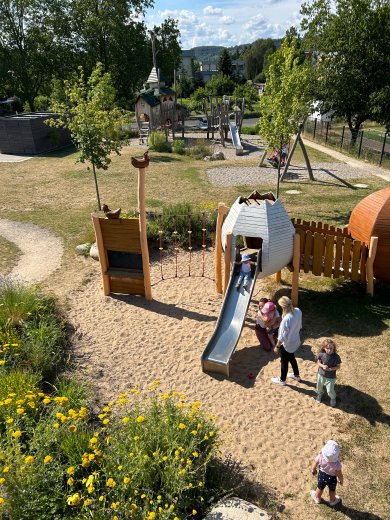 Ein naturnah gestalteter Spielbereich mit einer hölzernen Spielanlage in Form großer Eier und eines Hühnerstalls. Eine Edelstahlrutsche führt aus dem Spielgerät in den Sandbereich. Mehrere Kinder spielen gemeinsam an der Anlage, während eine pädagogische Fachkraft zwei Kinder begleitet. Im Vordergrund blühen gelbe Wildblumen, im Hintergrund befinden sich weitere Spielgeräte, Bäume und Grünflächen.