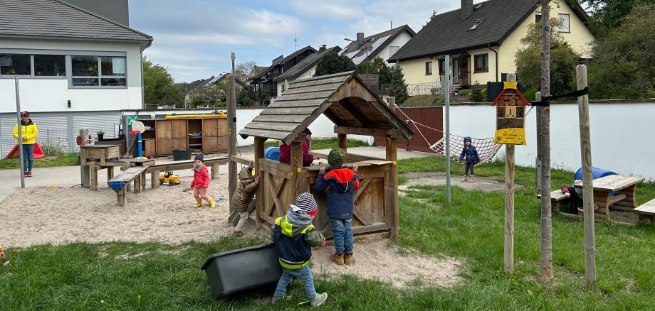 Kita Rambazamba Mehrere Kinder spielen auf dem Außengelände der Kindertagesstätte. Im Mittelpunkt steht ein kleines hölzernes Spielhäuschen, an dem Kinder gemeinsam spielen. Daneben befinden sich Sandflächen mit Spielmaterialien, eine Hängematte sowie weitere Spielstationen. Eine pädagogische Fachkraft steht am Rand des Geländes. Im Hintergrund sind Wohnhäuser, ein blühender Baum und die Gebäude der Kindertagesstätte zu sehen. Die naturnahe Außenanlage bietet vielfältige Bewegungs- und Spielmöglichkeiten.