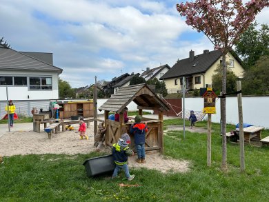 Mehrere Kinder spielen auf dem Außengelände der Kindertagesstätte. Im Mittelpunkt steht ein kleines hölzernes Spielhäuschen, an dem Kinder gemeinsam spielen. Daneben befinden sich Sandflächen mit Spielmaterialien, eine Hängematte sowie weitere Spielstationen. Eine pädagogische Fachkraft steht am Rand des Geländes. Im Hintergrund sind Wohnhäuser, ein blühender Baum und die Gebäude der Kindertagesstätte zu sehen. Die naturnahe Außenanlage bietet vielfältige Bewegungs- und Spielmöglichkeiten.