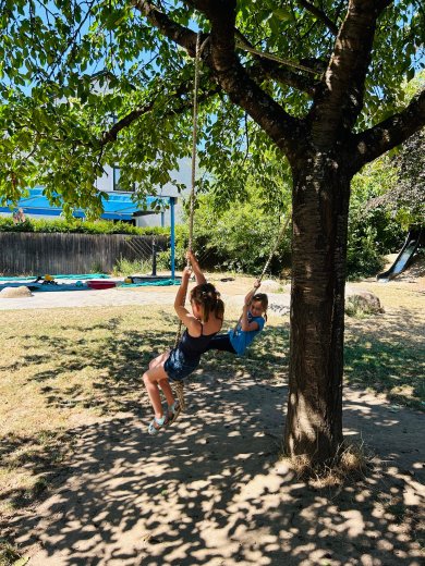 Zwei Kinder schwingen an Seilschaukeln, die an den kräftigen Ästen eines großen Laubbaums befestigt sind. Die Baumkrone spendet an einem sonnigen Sommertag angenehmen Schatten. Im Hintergrund sind weitere Bereiche des Außengeländes mit Sonnensegeln, Sandflächen und Spielgeräten zu erkennen. Die natürliche Umgebung lädt zum freien Bewegen und Spielen im Freien ein.
