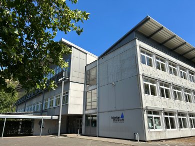 Schräg aufgenommene Außenansicht des Haupteingangs des Marion-Dönhoff-Gymnasiums. Das moderne Gebäude verfügt über eine helle Fassade mit großen Fensterfronten und einem markanten Dachüberstand. Über dem Eingang schützt ein Vordach vor Witterung. Der Gebäudename und das Schullogo sind an der Fassade angebracht. Die Architektur wirkt funktional und offen.