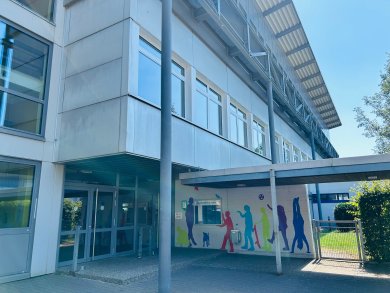 Der überdachte Haupteingang des Schulgebäudes mit Glas-Eingangstüren und breiten Stützpfeilern. An einer Seitenwand befinden sich farbenfrohe Silhouetten von Kindern in verschiedenen Bewegungs- und Spielsituationen. Der Eingangsbereich ist großzügig gestaltet und führt direkt in das Schulgebäude. Die moderne Architektur wird durch klare Linien und große Glasflächen geprägt