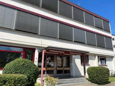Außenansicht eines modernen Schulgebäudes mit heller Fassade und großen Fenstern. Vor dem Haupteingang befindet sich ein überdachter Eingangsbereich mit roten Trägern und einer Treppe. Gepflegte Sträucher rahmen den Zugang ein. Die oberen Etagen verfügen über außenliegende Sonnenschutzlamellen, während der klare blaue Himmel die moderne Architektur des Gebäudes betont.