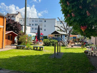 Großzügiges Außengelände einer Kindertagesstätte mit weitläufiger Rasenfläche, Bäumen und verschiedenen Spielbereichen. Links steht ein kleines Holzhaus, in der Mitte befinden sich Sitzbänke und ein Spielplatz mit Kletterturm, Rutsche und Spielhaus. Im Hintergrund sind weitere Spielgeräte und Wohnhäuser zu sehen. Ein großer Kastanienbaum spendet Schatten und ist mit bunten Dekorationen geschmückt.