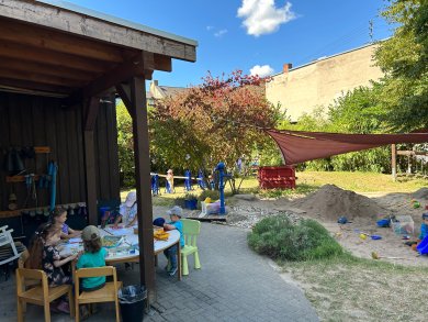 Mehrere Kinder sitzen unter einem überdachten Holzunterstand an einem runden Tisch und malen oder basteln gemeinsam. Rechts schließt sich ein großer Sandspielbereich mit Sandhügel, Spielzeug und einer Wasserpumpe an. Ein Sonnensegel spendet Schatten. Sträucher, Bäume und weitere Spielbereiche schaffen eine naturnahe Umgebung.