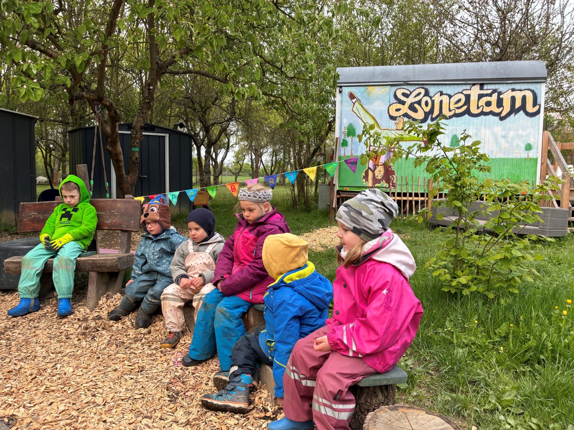 Sechs Kinder in wetterfester, farbenfroher Kleidung sitzen nebeneinander auf Holzbänken und Baumstämmen auf dem Gelände eines Waldkindergartens. Im Hintergrund stehen Bäume und dunkle Holzhütten sowie ein bunt bemalter Bauwagen mit der großen Aufschrift „Lahnstein“. Eine Wimpelkette mit der Aufschrift „Happy Birthday“ ist zwischen den Bäumen gespannt. Der Boden ist teilweise mit Holzspänen bedeckt, rund um den Sitzbereich wachsen Gras und Sträucher.
