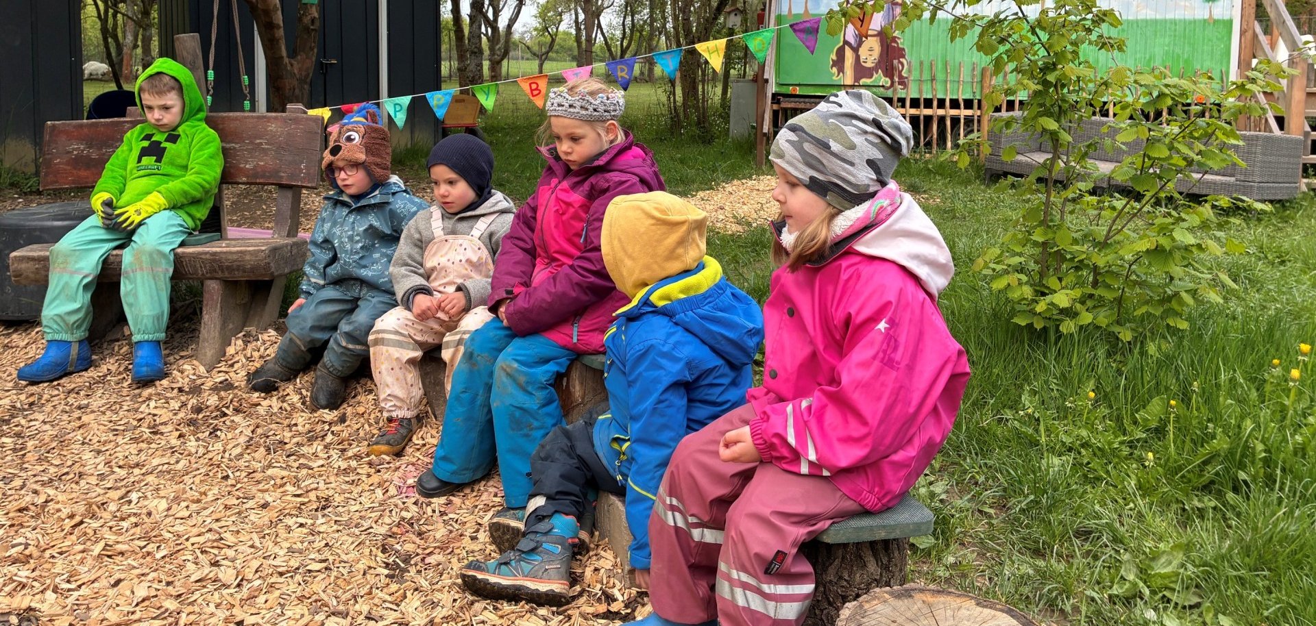 Sechs Kinder in wetterfester, farbenfroher Kleidung sitzen nebeneinander auf Holzbänken und Baumstämmen auf dem Gelände eines Waldkindergartens. Im Hintergrund stehen Bäume und dunkle Holzhütten sowie ein bunt bemalter Bauwagen mit der großen Aufschrift „Lahnstein“. Eine Wimpelkette mit der Aufschrift „Happy Birthday“ ist zwischen den Bäumen gespannt. Der Boden ist teilweise mit Holzspänen bedeckt, rund um den Sitzbereich wachsen Gras und Sträucher.