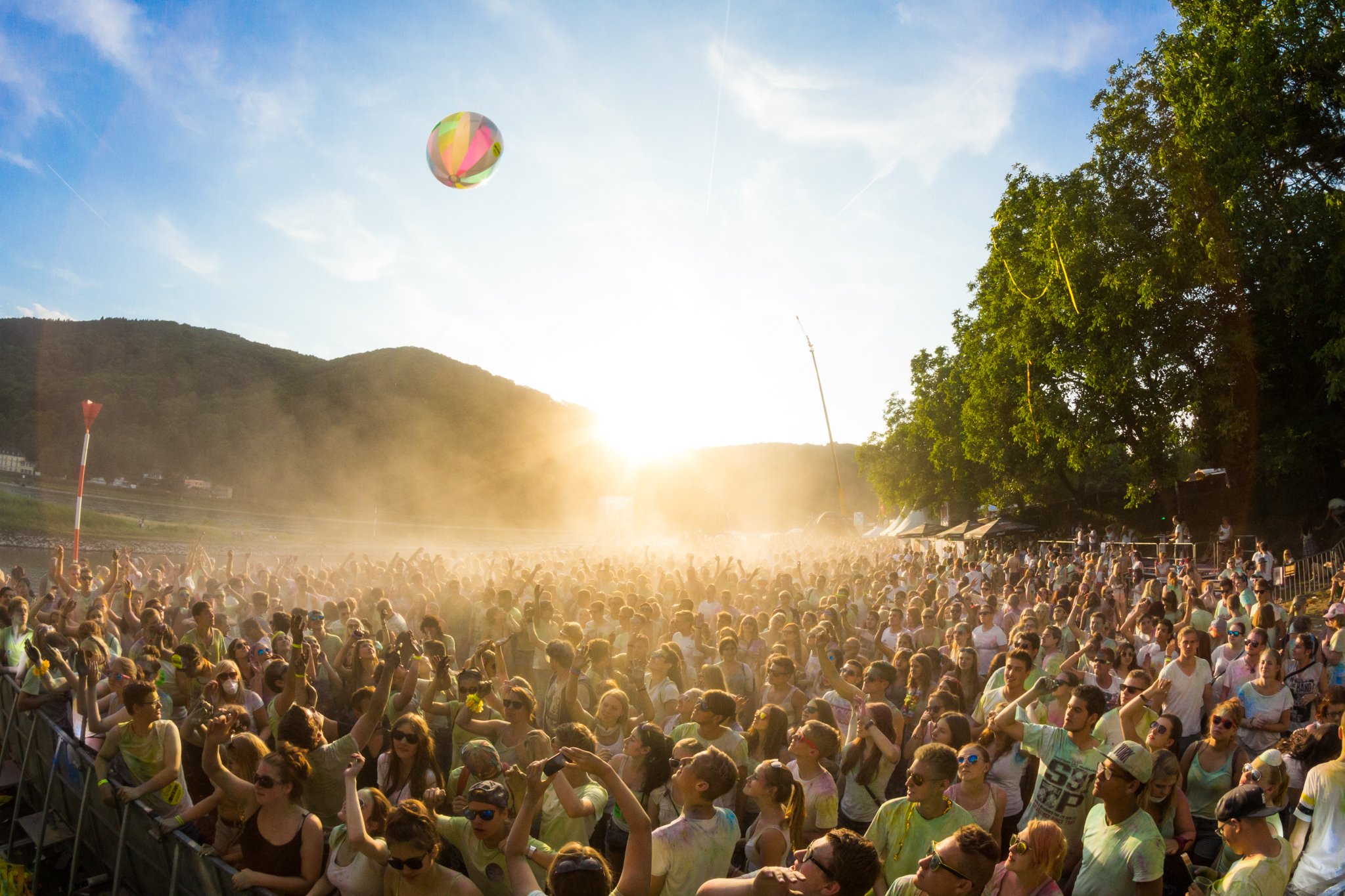 Große Menschenmenge bei einer Open-Air-Veranstaltung im Freien. Viele Besucherinnen und Besucher stehen dicht beieinander und strecken die Arme in die Höhe. Über der Menge fliegt ein bunter Strandball. Im Gegenlicht der tief stehenden Sonne liegt feiner Staub oder Farbpulver in der Luft, im Hintergrund sind bewaldete Hügel zu sehen.