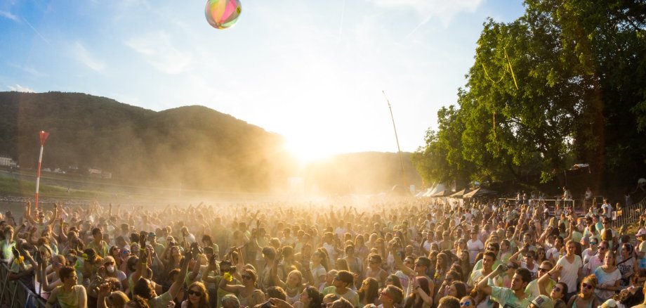 Veranstaltungen Lahnstein Große Menschenmenge bei einer Open-Air-Veranstaltung im Freien. Viele Besucherinnen und Besucher stehen dicht beieinander und strecken die Arme in die Höhe. Über der Menge fliegt ein bunter Strandball. Im Gegenlicht der tief stehenden Sonne liegt feiner Staub oder Farbpulver in der Luft, im Hintergrund sind bewaldete Hügel zu sehen.