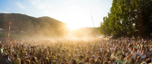 Veranstaltungen Lahnstein Große Menschenmenge bei einer Open-Air-Veranstaltung im Freien. Viele Besucherinnen und Besucher stehen dicht beieinander und strecken die Arme in die Höhe. Über der Menge fliegt ein bunter Strandball. Im Gegenlicht der tief stehenden Sonne liegt feiner Staub oder Farbpulver in der Luft, im Hintergrund sind bewaldete Hügel zu sehen.