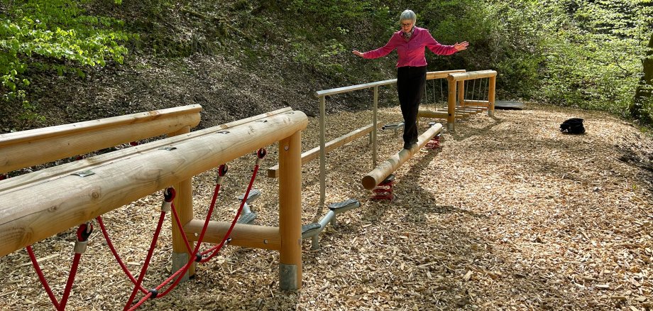 Eine Frau balanciert mit ausgestreckten Armen über einen schmalen Holzstamm eines Bewegungsparcours im Wald. Die Station ist von Holzschnitzeln umgeben und liegt inmitten eines dichten Laubwaldes mit frischem Grün.