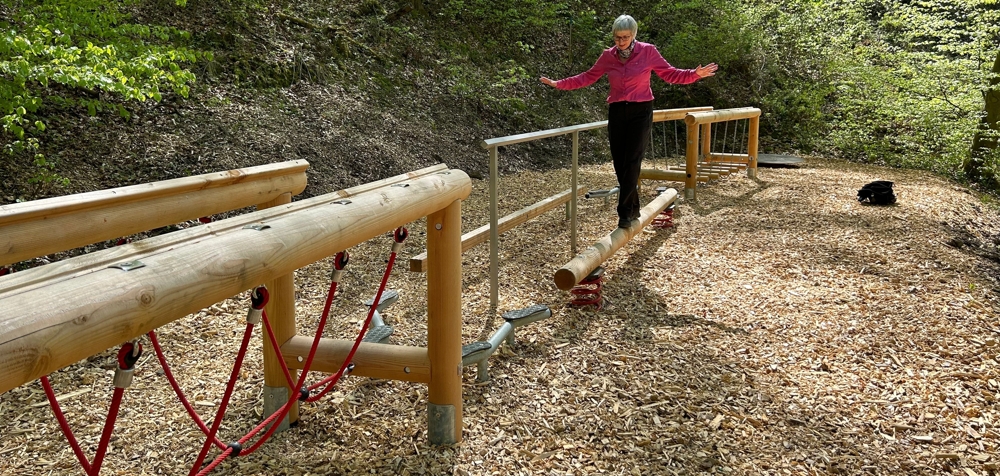 Eine Frau balanciert mit ausgestreckten Armen über einen schmalen Holzstamm eines Bewegungsparcours im Wald. Die Station ist von Holzschnitzeln umgeben und liegt inmitten eines dichten Laubwaldes mit frischem Grün.