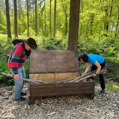 Atemparcours Kur- und Heilwald Lahnstein Zwei Personen öffnen gemeinsam eine große Holzkiste an einer Station im Wald und entnehmen lange Holzstäbe. Die Station steht auf einer mit Holzschnitzeln bedeckten Fläche und ist von hohen Bäumen, dichtem Grün und Unterholz umgeben.