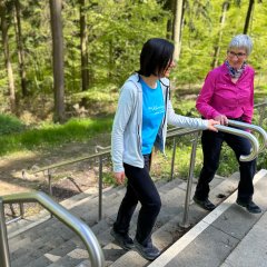Atemparcours Kur- und Heilwald Lahnstein Zwei Frauen gehen gemeinsam eine Treppe im Kur- und Heilwald hinauf und halten sich dabei am Handlauf fest. Im Hintergrund sind ein Waldweg und dichtes Frühlingsgrün zu sehen, die die naturnahe Umgebung des Bewegungsparcours unterstreichen.