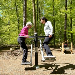 Zwei Frauen nutzen ein Fitnessgerät im Wald. Sie stehen sich auf gegenüberliegenden Trittflächen eines Trainingsgeräts und halten sich an den Griffstangen fest. Die Outdoor-Fitnessstation steht auf einem befestigten Platz, umgeben von dichtem grünem Wald mit hohen Bäumen und sonnigem Licht.