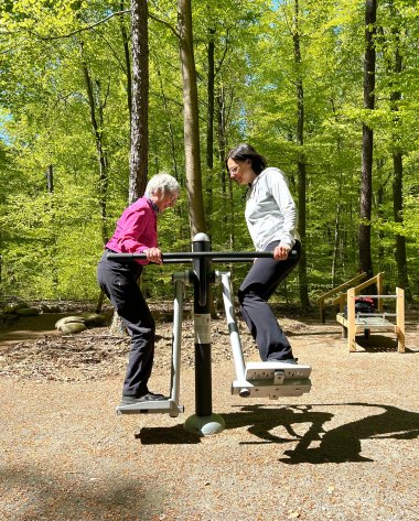 Zwei Frauen nutzen ein Fitnessgerät im Wald. Sie stehen sich auf gegenüberliegenden Trittflächen eines Trainingsgeräts und halten sich an den Griffstangen fest. Die Outdoor-Fitnessstation steht auf einem befestigten Platz, umgeben von dichtem grünem Wald mit hohen Bäumen und sonnigem Licht.
